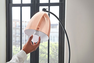   A smart light bulb held by a hand and being screwed on the lamp socket of a table lamp 